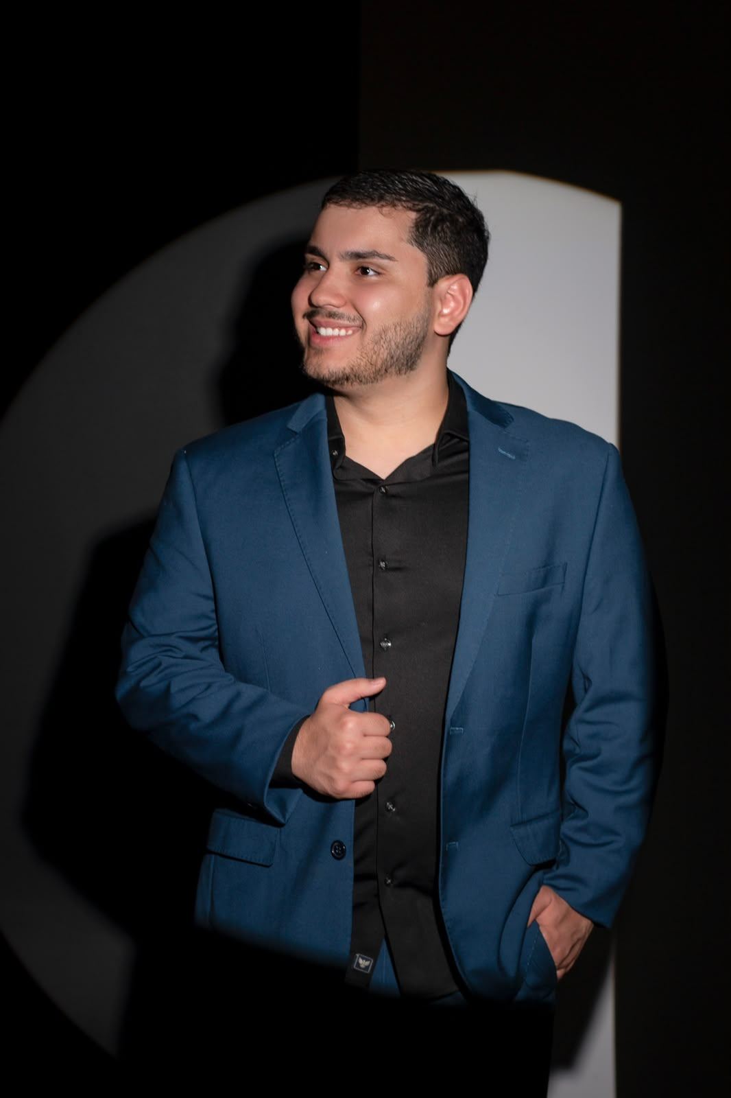Marcus Trivellato de pé, sorrindo, de blazer azul-marinho e camisa preta